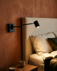 Arbotante LED de lectura BRACCIO negro con brazo articulado encendido junto a cabecera de lino en recámara de muro terracota