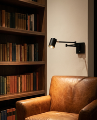 Lámpara de pared BRACCIO con brazo extendido sobre sillón de piel cognac junto a librero de nogal