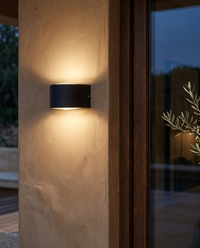 Lámpara de pared exterior CINCEL con luz cálida en muro de estuco junto a puerta de terraza de madera oscura