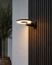 Lámpara de pared exterior DISCOR WALL con disco de luz cálida en terraza de estuco blanco con lambrín de madera oscura y olivo
