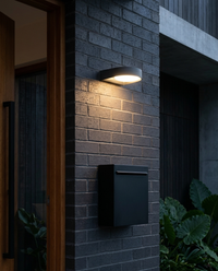 Arbotante LED GIDI gris oscuro encendido en fachada de ladrillo carbón sobre buzón negro junto a la puerta de teca